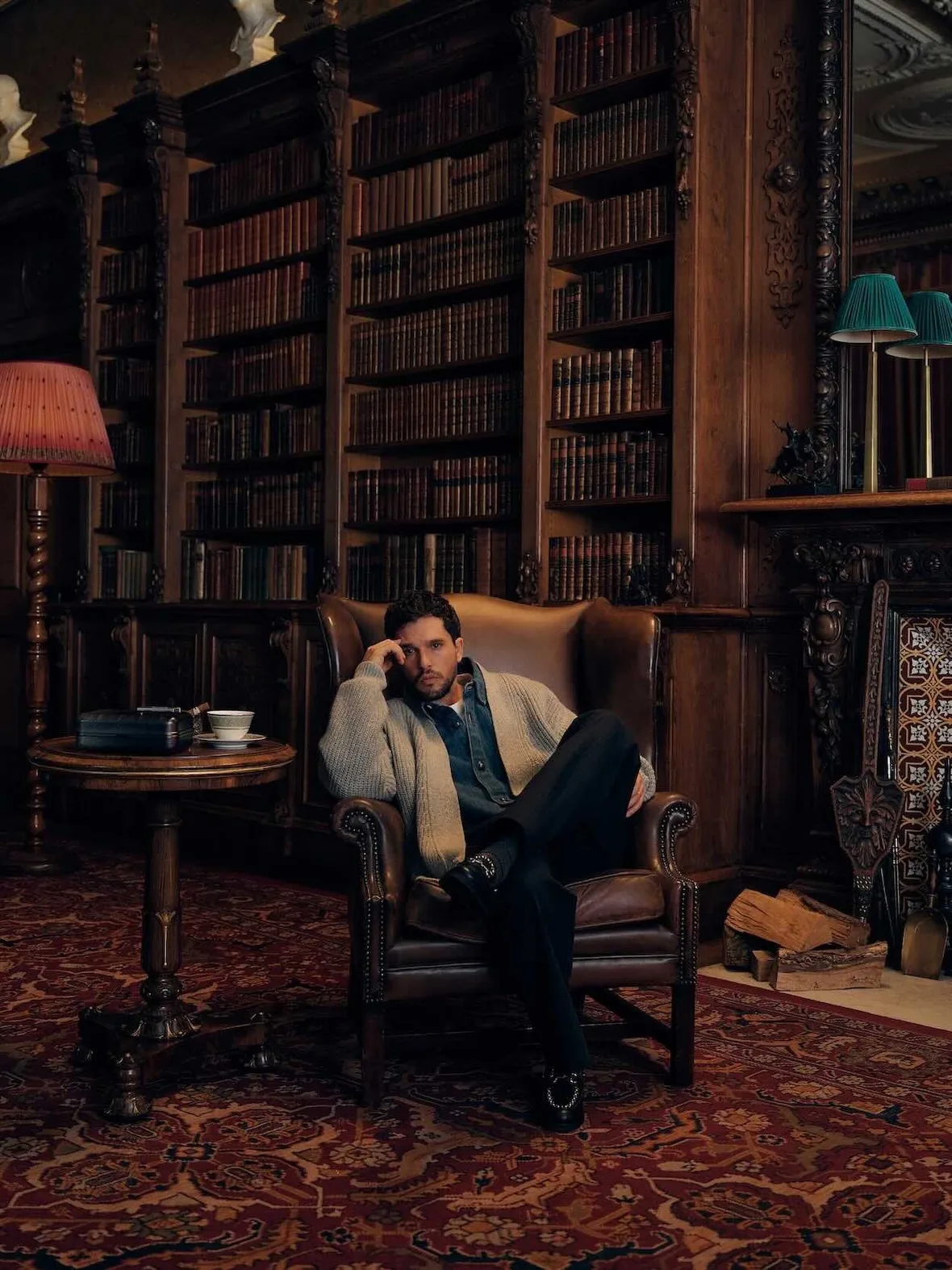On-model, seated in brown leather wingback chair in wood-panelled library, beige cardigan, black loafers, red rug, Winter