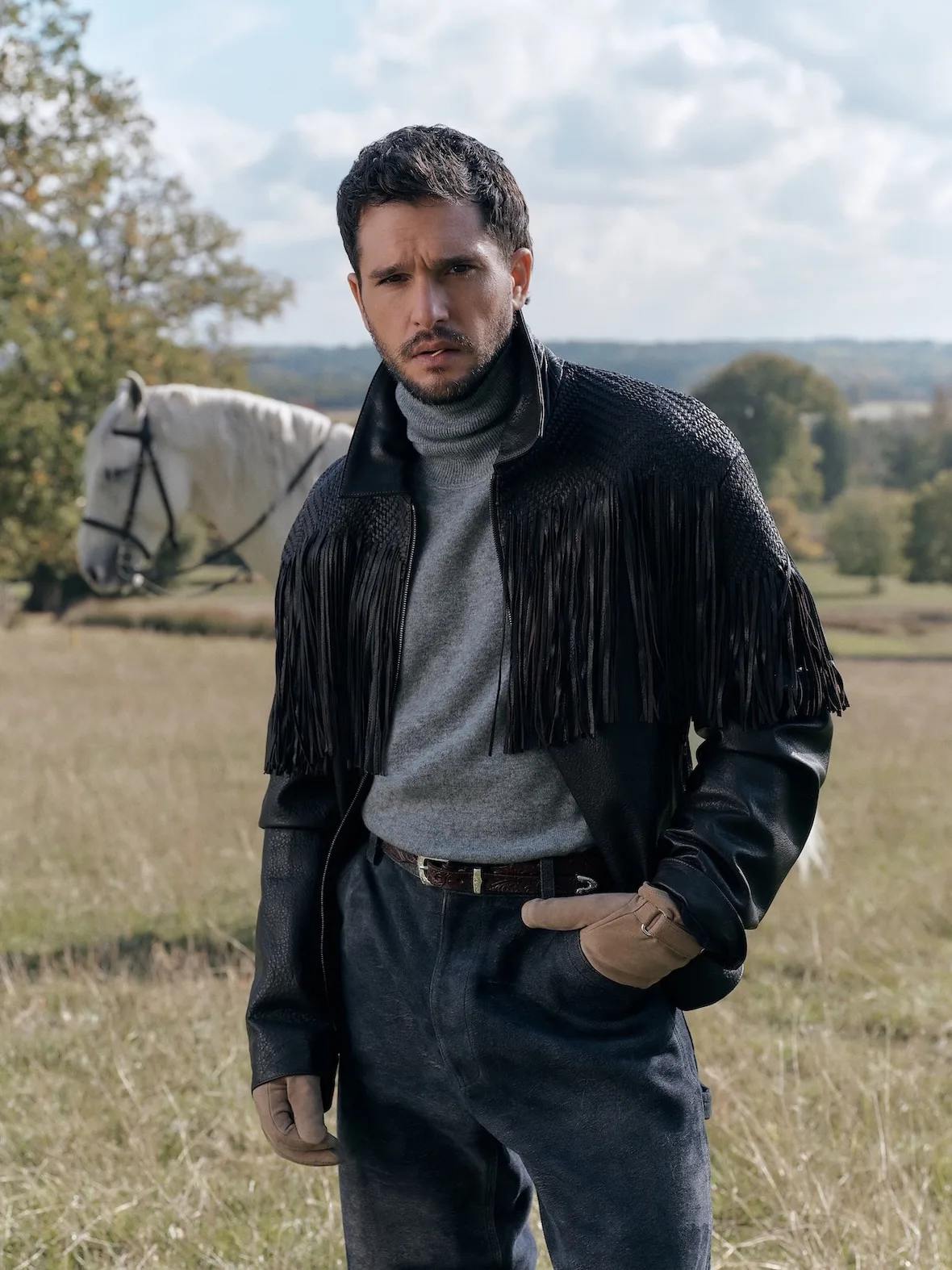 On-model, standing in field, black woven fringed leather jacket, grey turtleneck, tan gloves, white horse behind, Winter