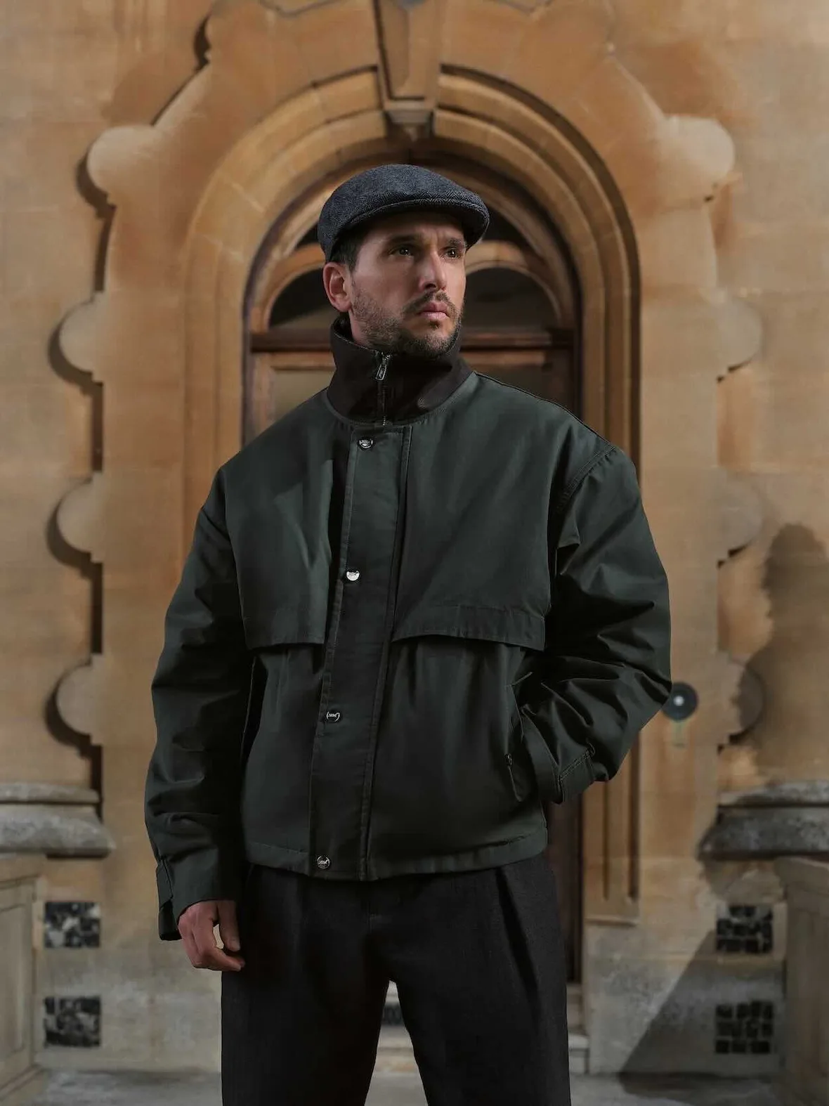 On-model, standing before stone arched doorway, dark green zip jacket with snap buttons, grey flat cap, Winter 2026