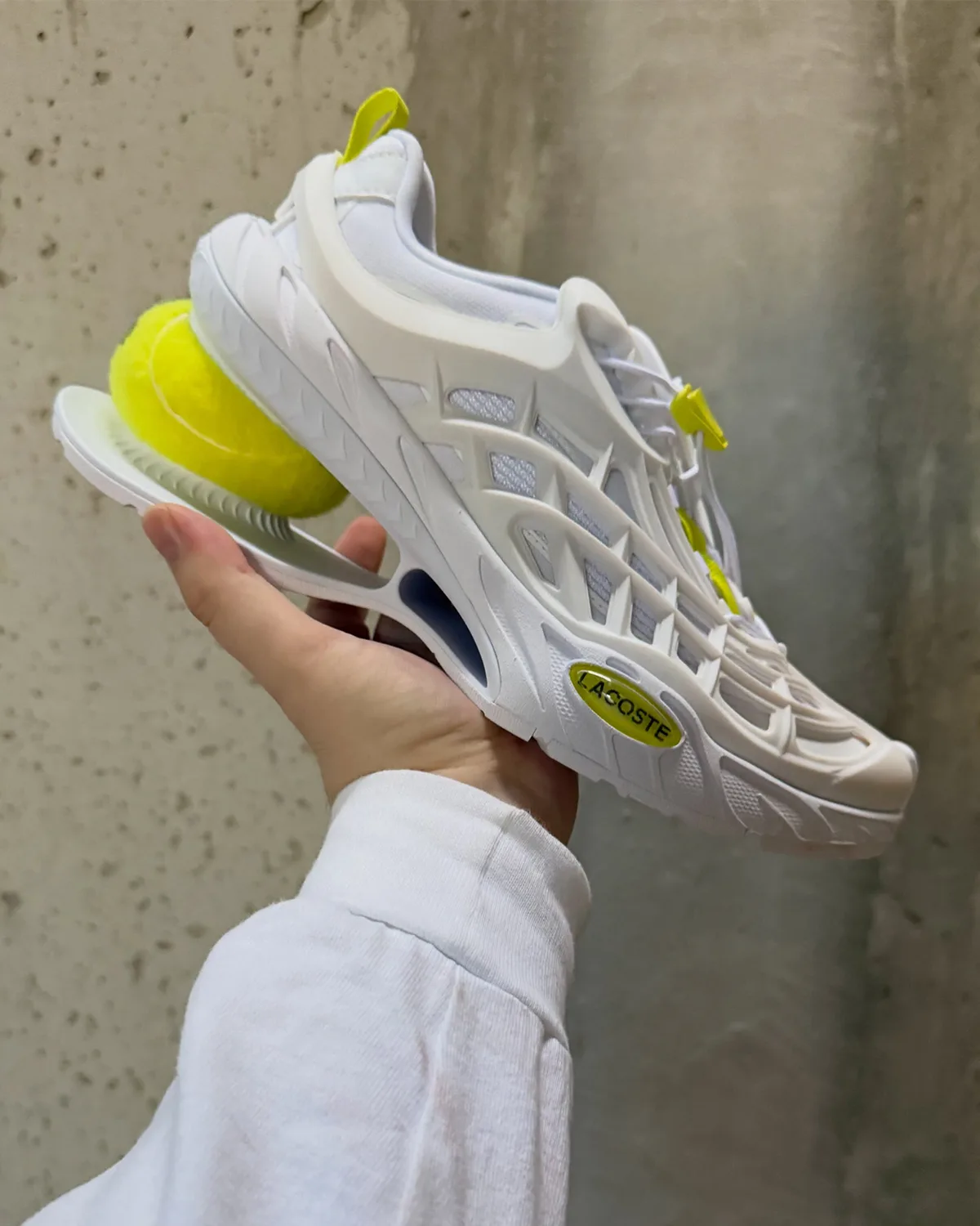 In-hand lateral view of a white shoe with a tennis ball heel, yellow-green toggles and oval Lacoste badge, Lacoste Ace T