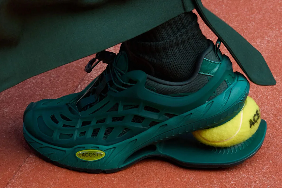 On-foot lateral view of a dark green caged shoe with a tennis ball heel and yellow Lacoste oval badge, Lacoste Ace T