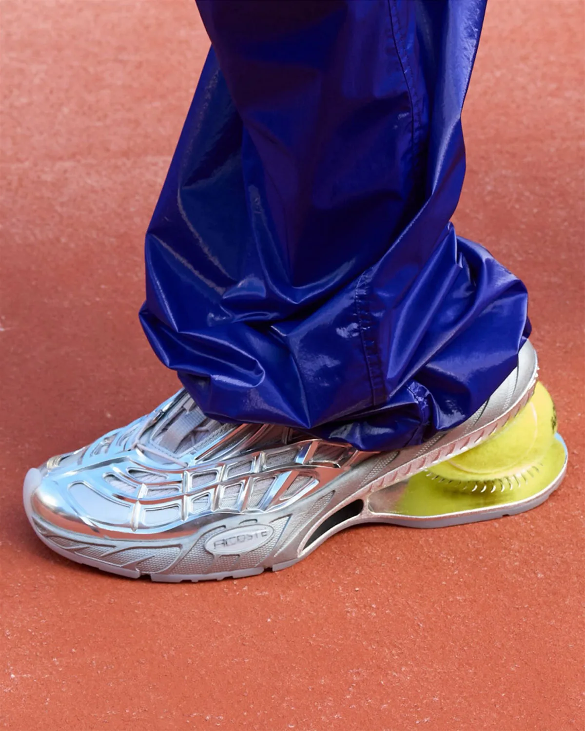 On-foot lateral view of metallic silver shoe with a tennis ball heel, shiny blue trousers, clay court, Lacoste Ace T