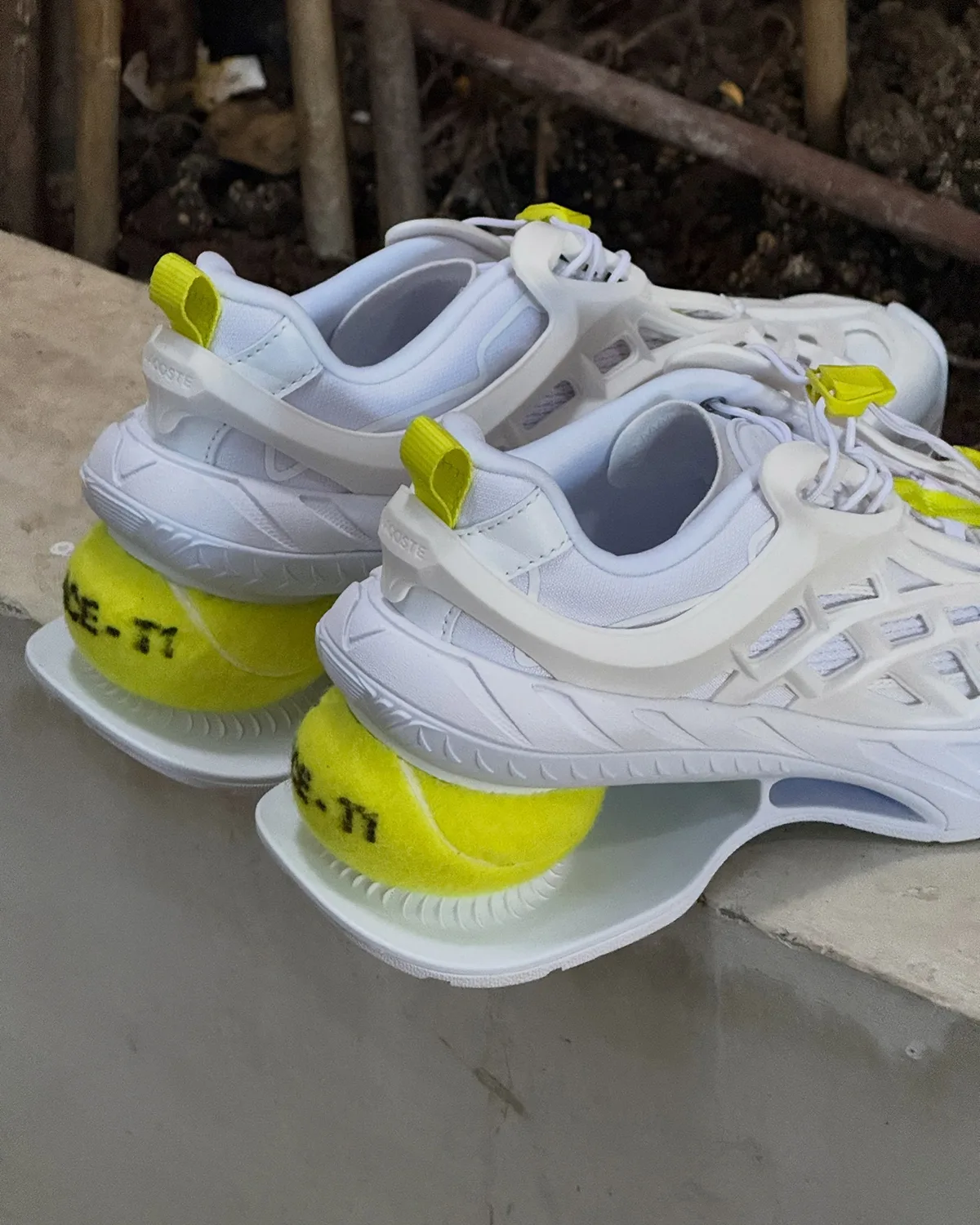 Pair of white shoes on a concrete ledge, each with a tennis ball heel and yellow-green pull tab, Lacoste Ace T