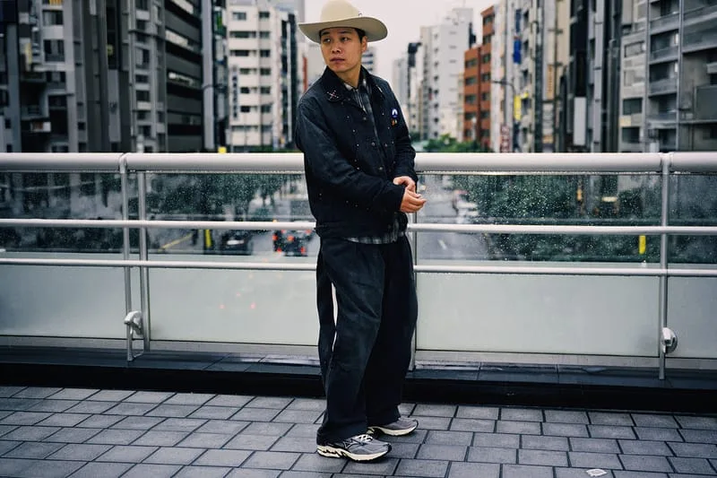 Person in cream cowboy hat and dark denim outfit on a city footbridge, wearing grey striped sneakers, Sean Wotherspoon Pack