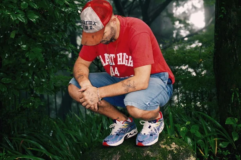 Person in red cap and tee crouching on a mossy rock in greenery, blue striped sneakers visible, Sean Wotherspoon Pack