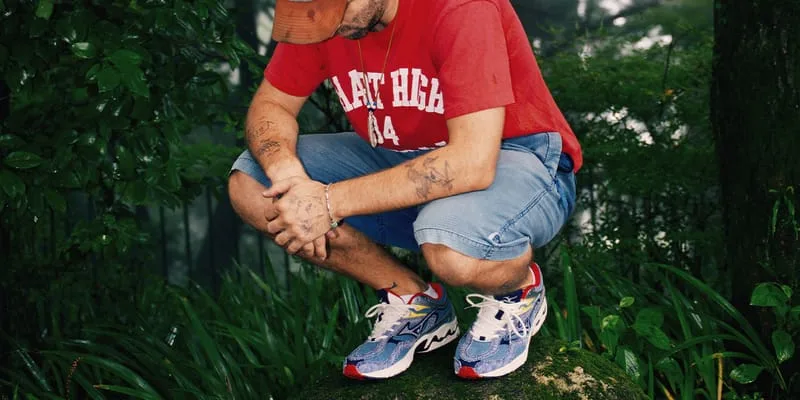 Person crouching on a mossy rock in a forest, wearing blue striped sneakers, red tee and denim shorts, Sean Wotherspoon Pack