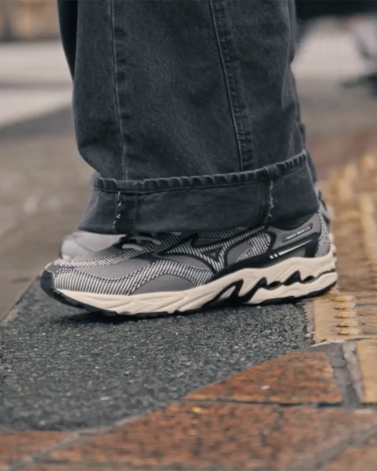 Lateral on-foot view of grey and black striped sneakers with cream wavy midsole, cuffed jeans, tactile paving, Sean