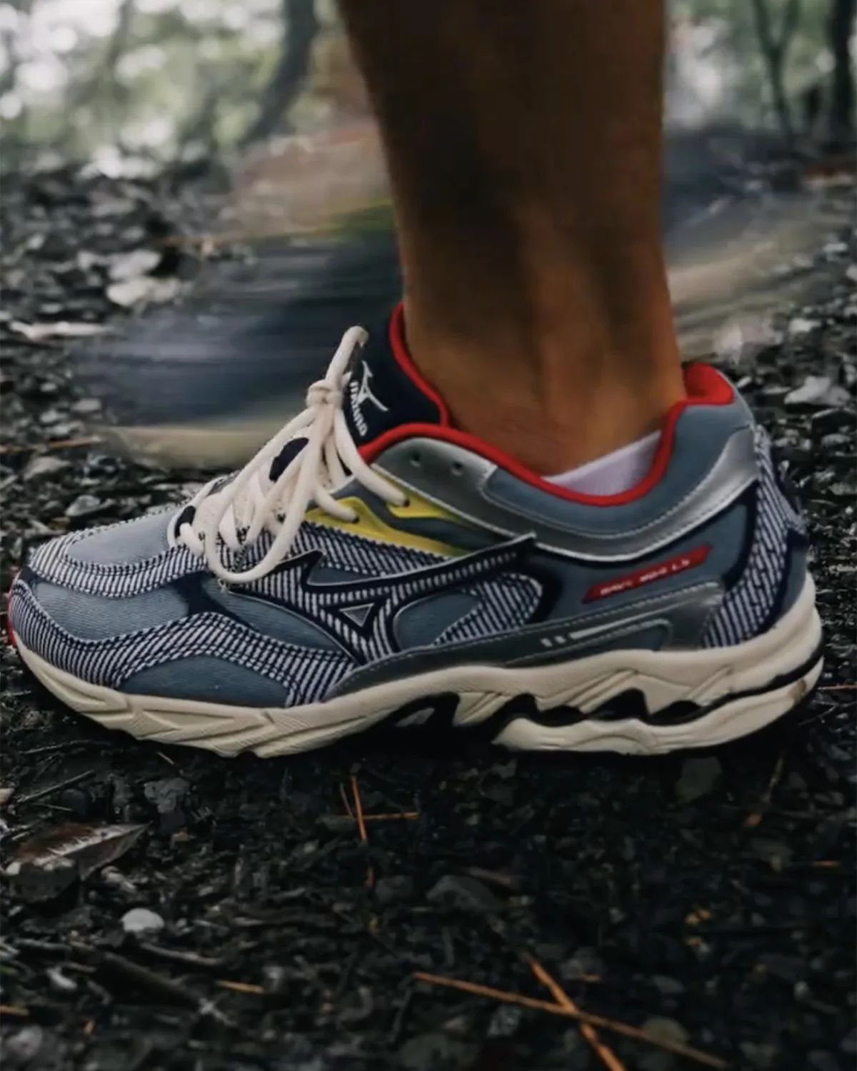Lateral on-foot view of blue striped sneaker with red collar, yellow accent and cream laces on dark gravel, Sean Wotherspoon