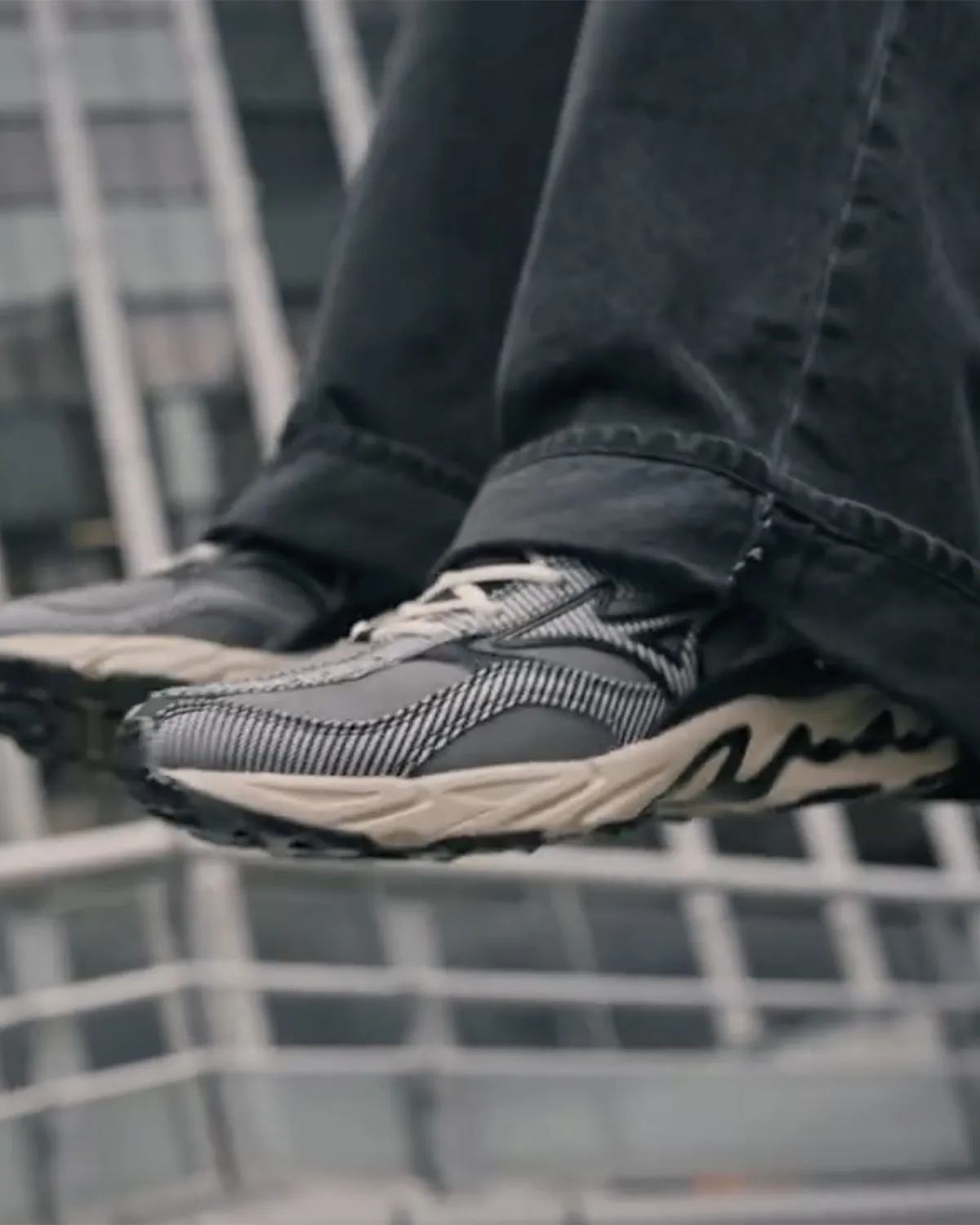 On-foot view of grey striped sneaker with cream wavy sole, jeans cuffed, blurred railing behind, Sean Wotherspoon Pack