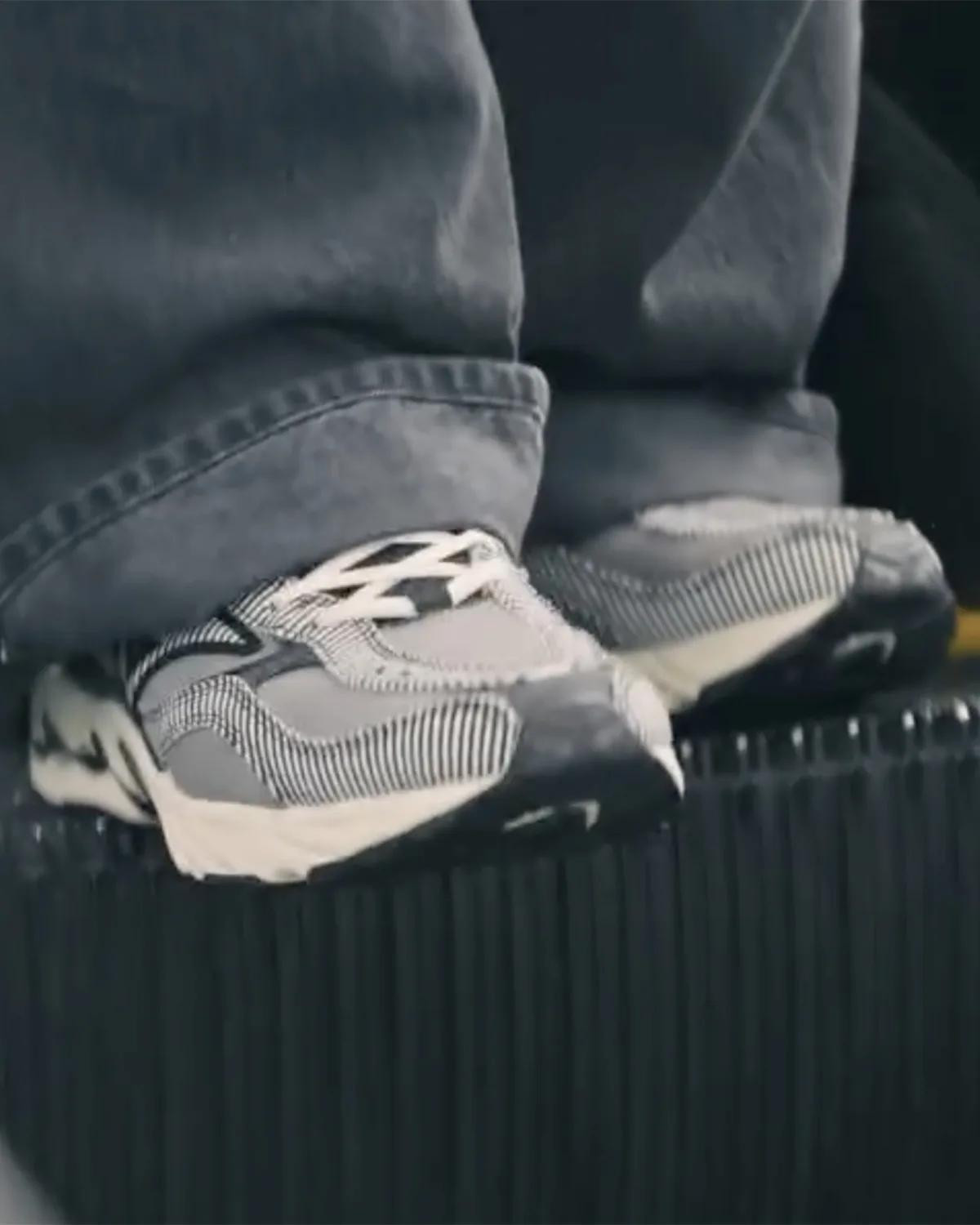 On-foot close-up of grey and white striped sneakers under baggy grey jeans, on a ribbed black surface, Sean Wotherspoon Pack