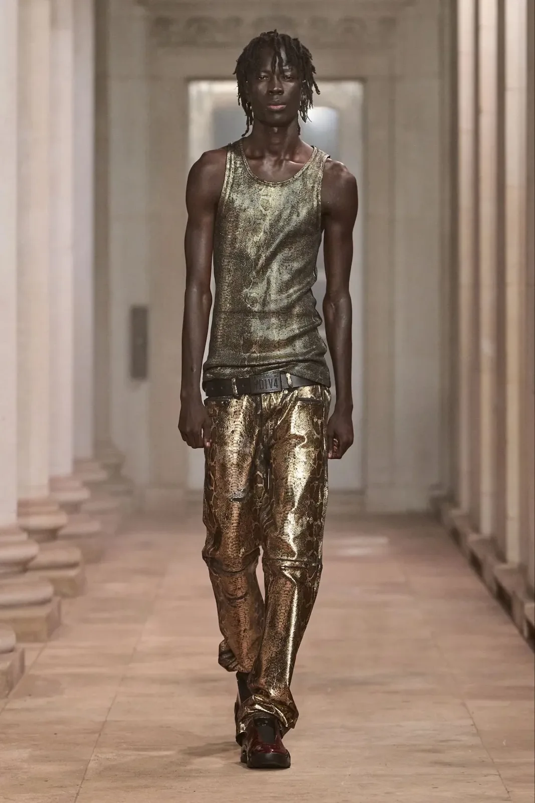 Runway model in gold metallic snakeskin-print trousers and tank top, dark red shoes at the feet, Patent Croc