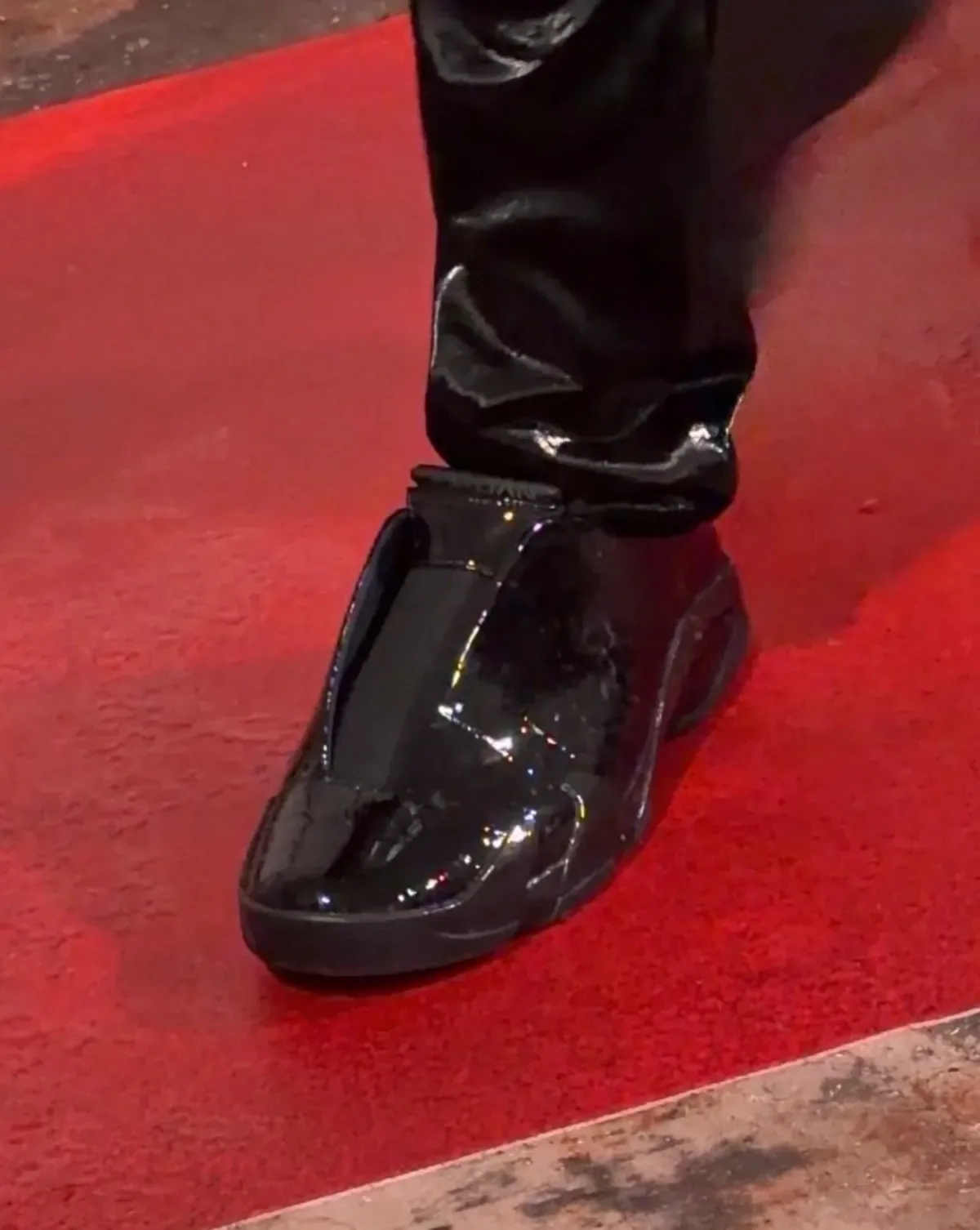 On-foot close-up of all-black patent mule on a red carpet, shiny black trouser hem, Patent Croc