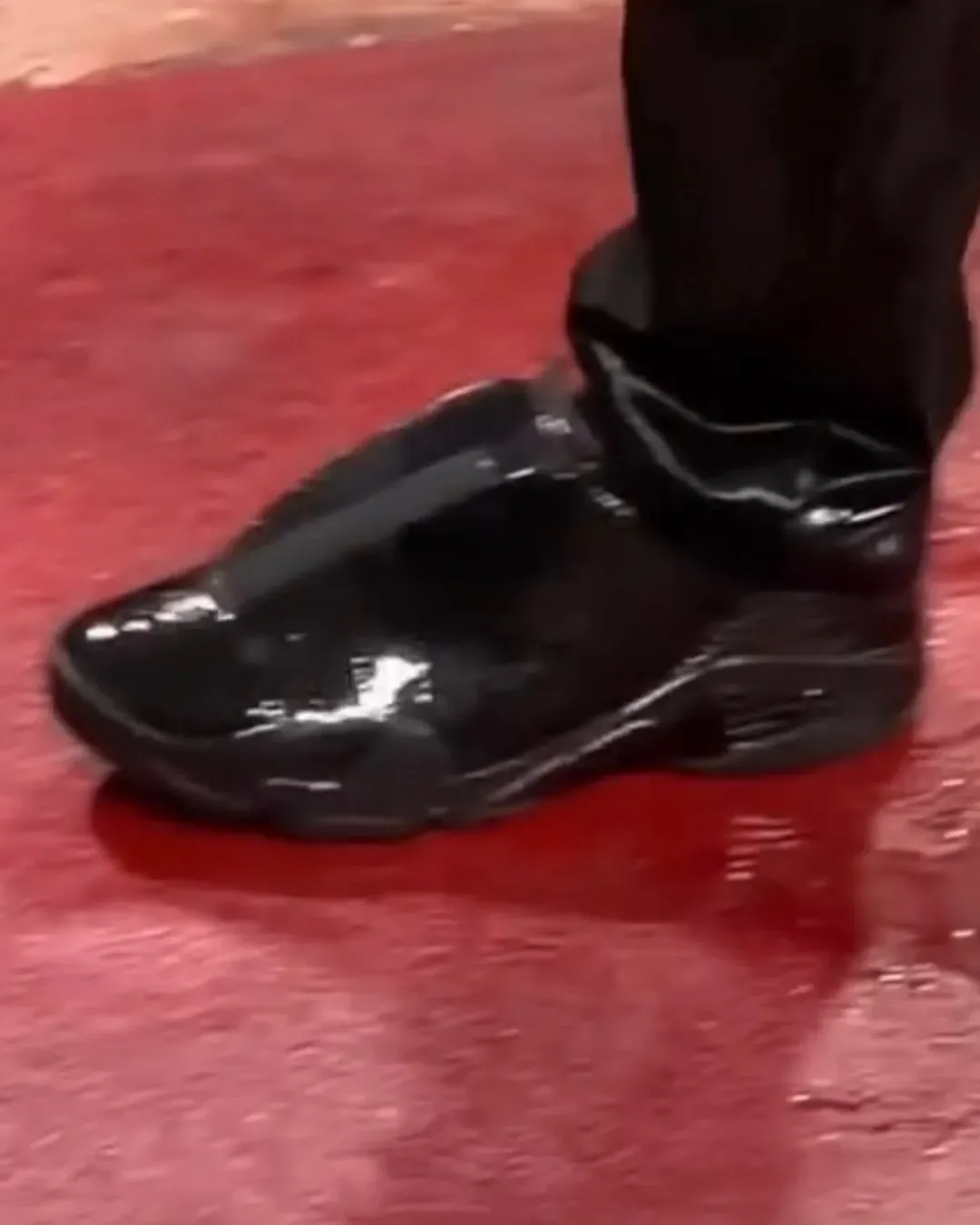 Lateral on-foot view of glossy black patent mule on red floor, black ribbed trouser leg, Patent Croc