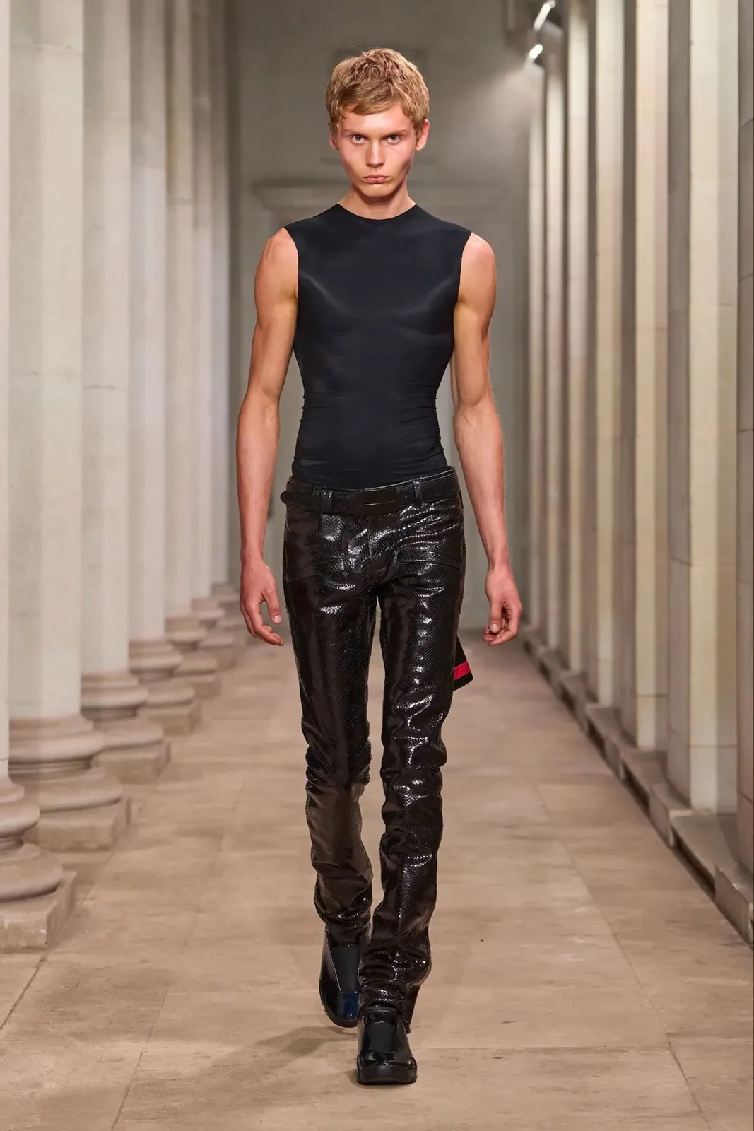 Runway model walking a stone colonnade in black snakeskin-look trousers and black glossy shoes, Patent Croc