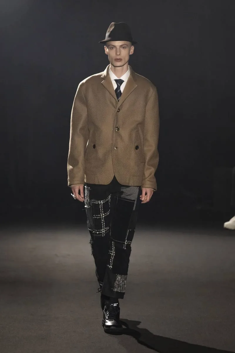 Runway model in a brown blazer, fedora and patchwork black trousers, wearing dark sneakers, Watanabe MAN P400