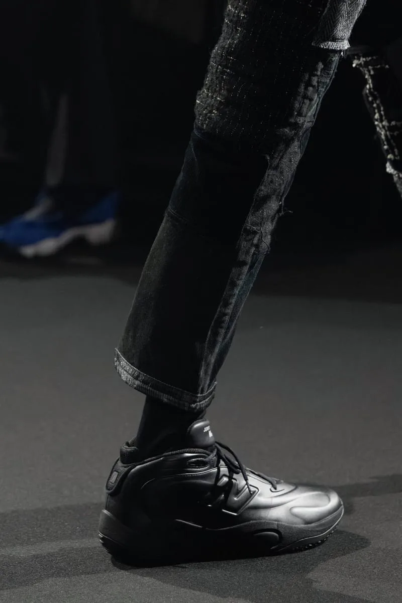 On-foot runway detail of a black sneaker under rolled, patchwork dark trousers on a dark stage, Watanabe MAN P400