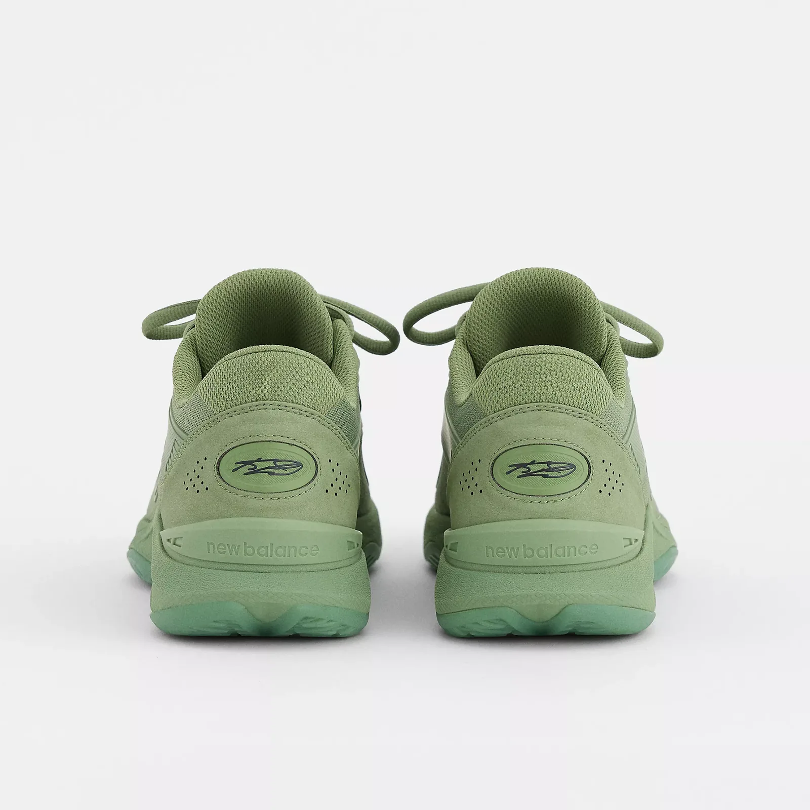 Heel view of pair in tonal green suede, oval signature logos and new balance text on heel, Oregano