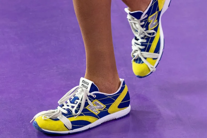 On-foot lateral view of blue and yellow sneaker, miu miu heel tab, grey N logo, raised foot behind, New Balance RC150