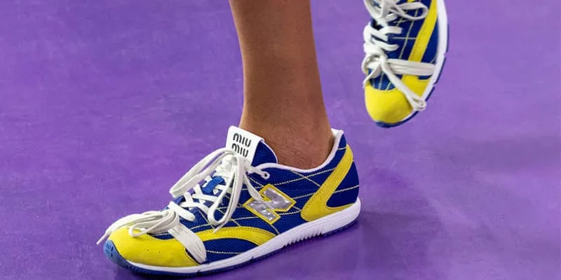 On-foot lateral view of blue and yellow sneaker with white laces and miu miu heel tab on purple floor, New Balance RC150