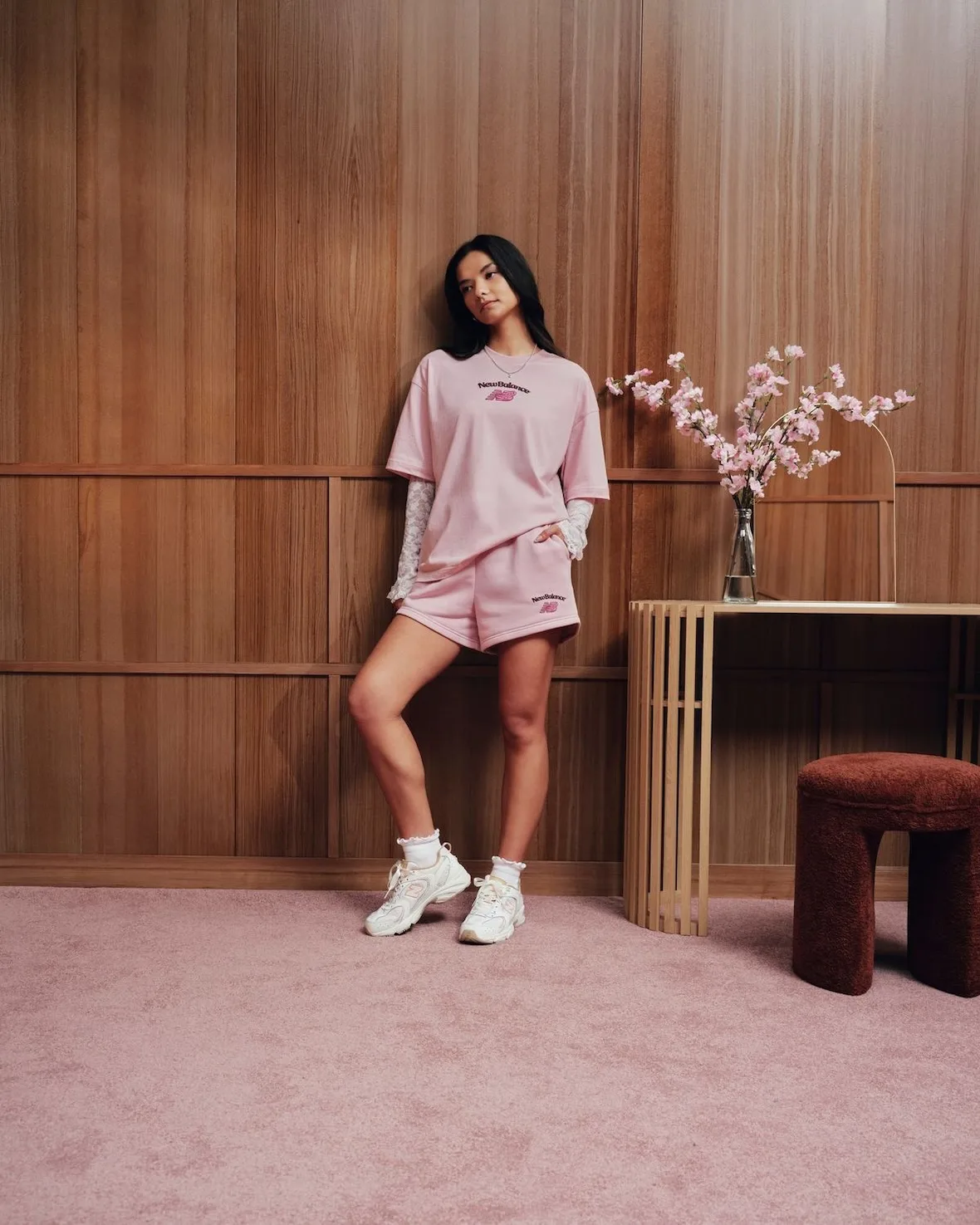 Model in pink tee and shorts with white sneakers, blossom vase and rust stool nearby, Sakura