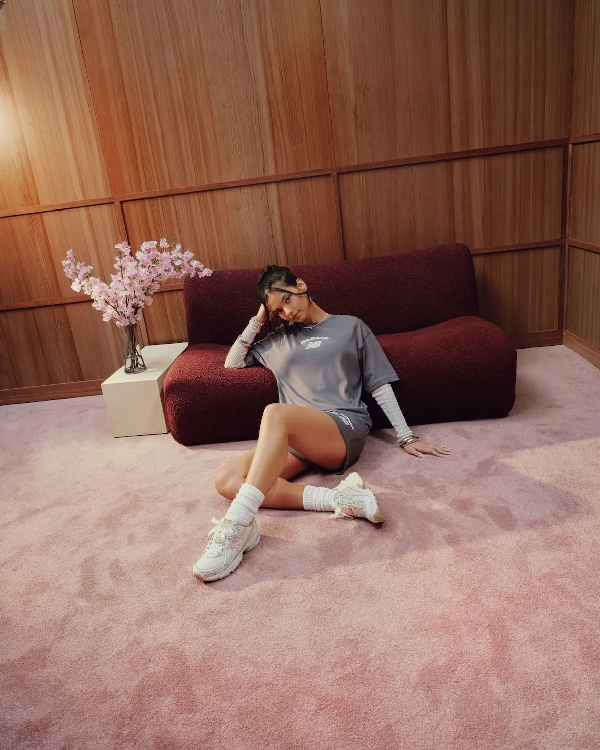 Model seated on pink carpet against burgundy sofa in grey outfit, cream sneakers, slouch socks, Sakura