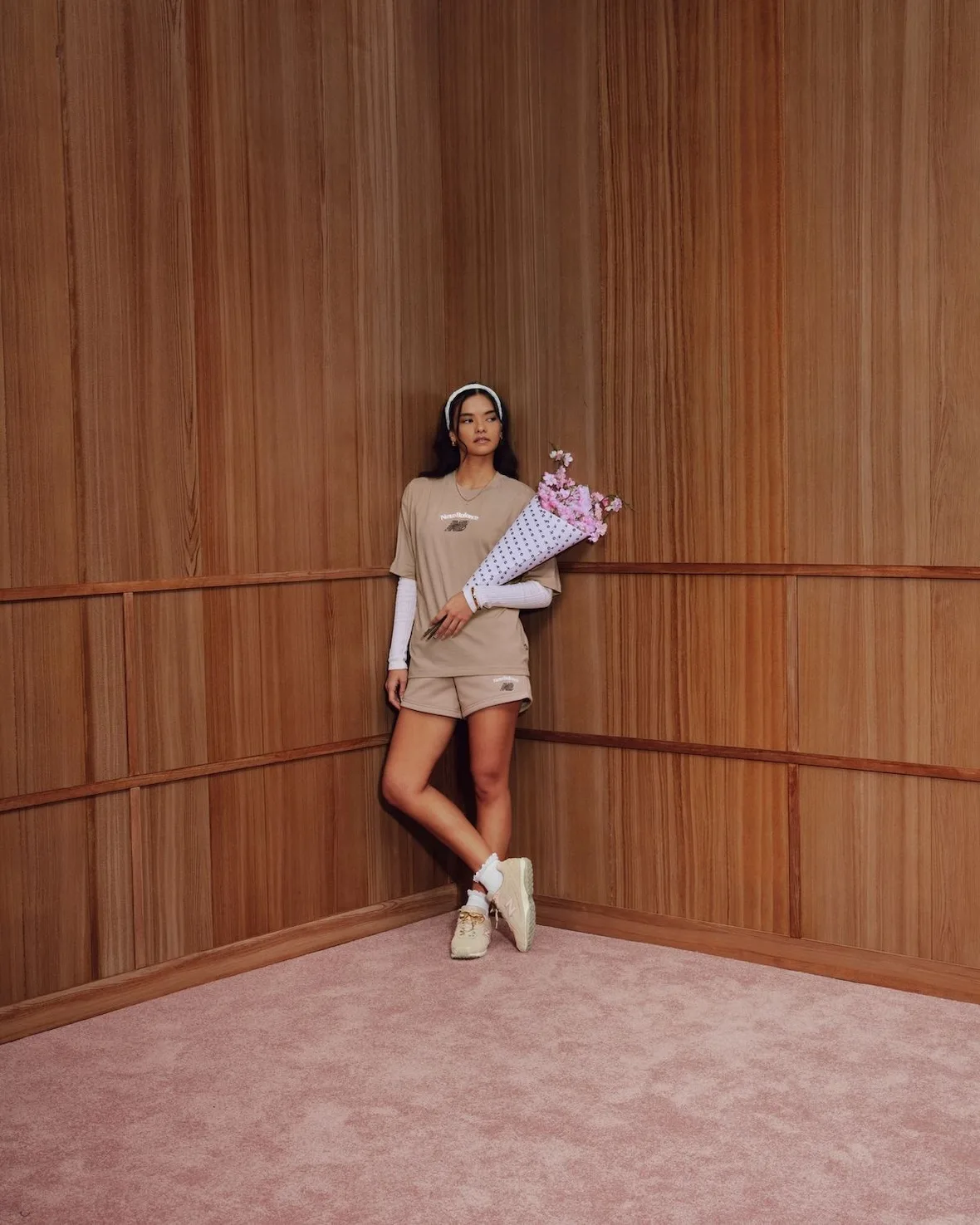 Model leaning in wood-panelled corner in tan outfit with blossom bouquet, tan sneakers on feet, Sakura