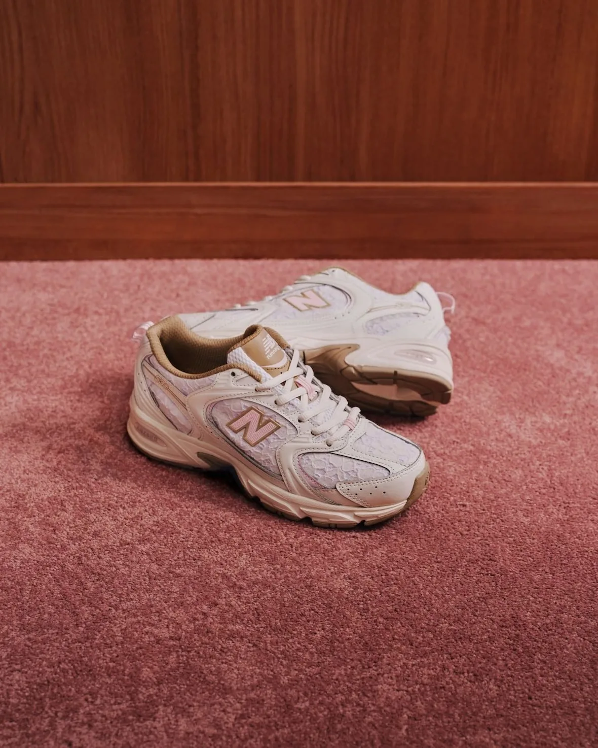 Lateral view of cream lace sneaker pair with pink N logos on pink carpet by wood skirting, Sakura