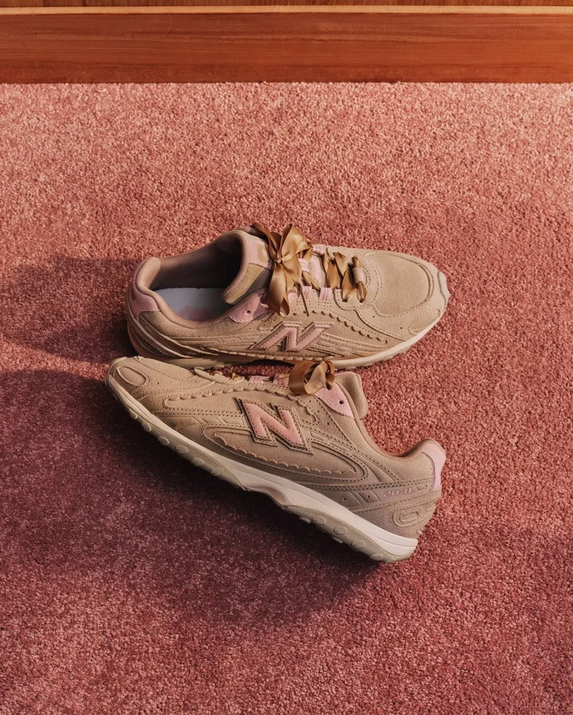 Pair of tan suede sneakers with gold ribbon laces and pink N logos on rust carpet, Sakura