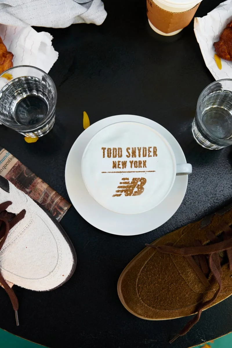 Top-down cafe table with Todd Snyder New York latte art, cream and brown suede sneakers at edges, Café