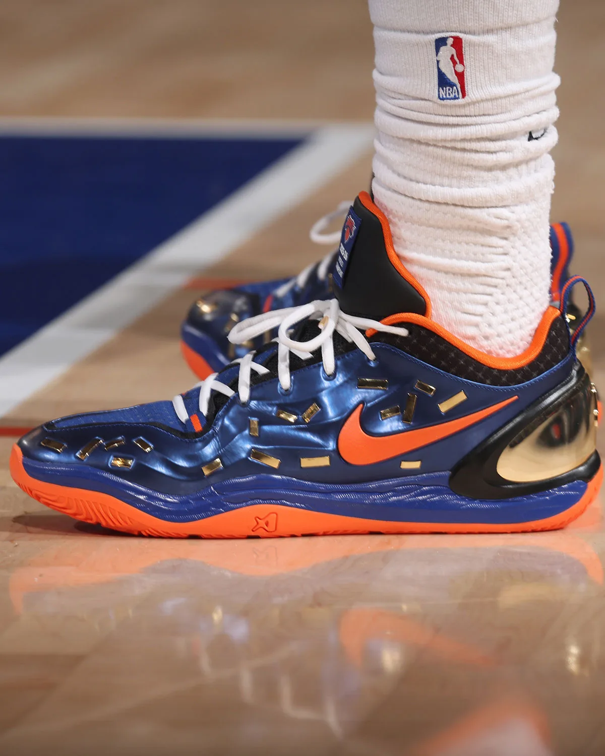 On-foot lateral view on hardwood: glossy blue upper with orange Swoosh, scattered gold chips, orange outsole, Confetti