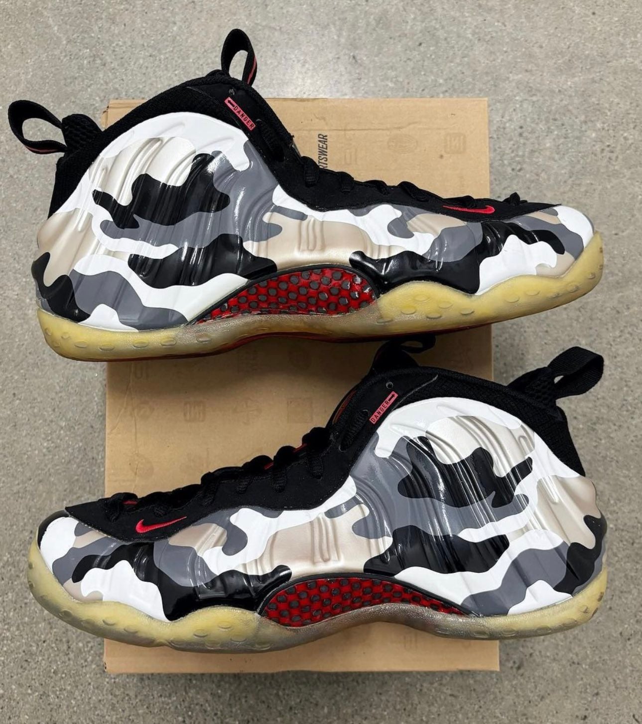 Top-down pair on a cardboard box on concrete, camo shell, red swoosh, yellowed translucent soles, Fighter Jet