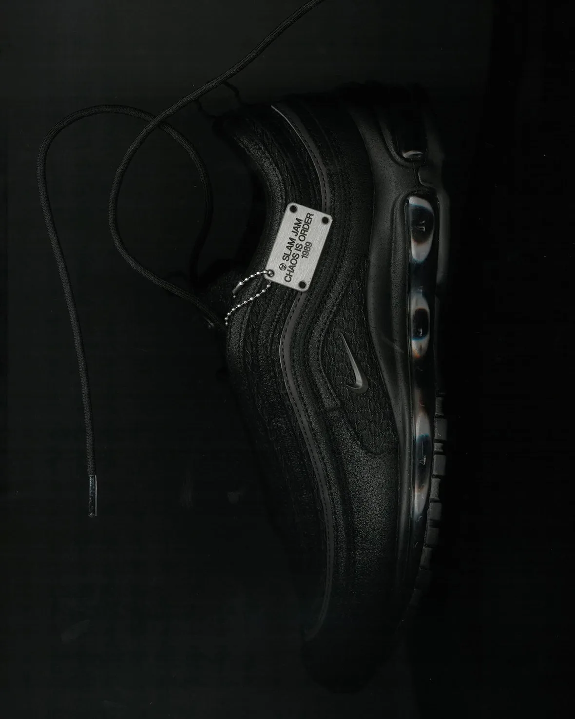 Top-down view of black shoe on dark ground with loose lace and silver Slam Jam tag, Black
