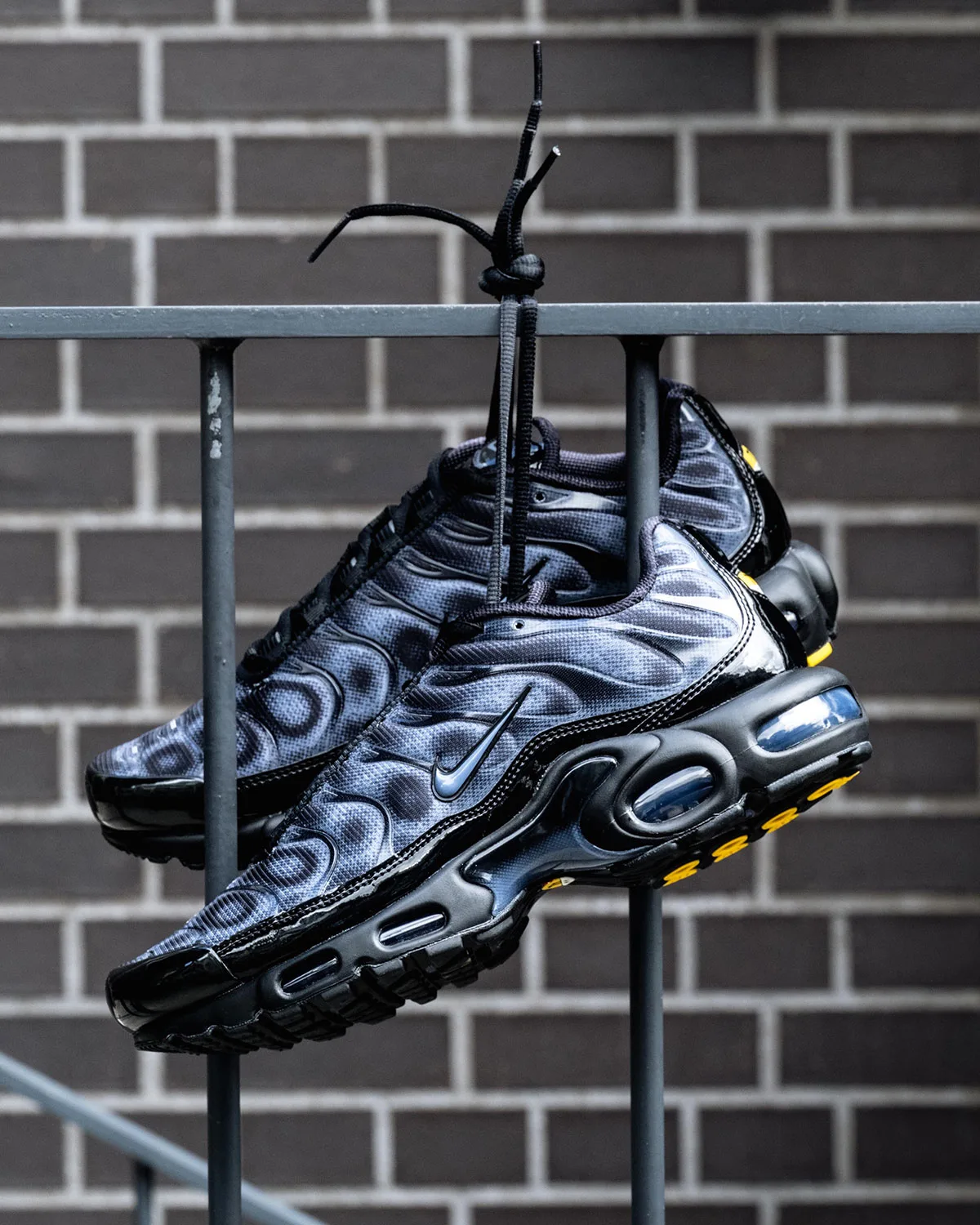 Lateral view of pair hanging from railing by laces, yellow tread and blue-tinted air unit, brick wall, Black/Cool Grey