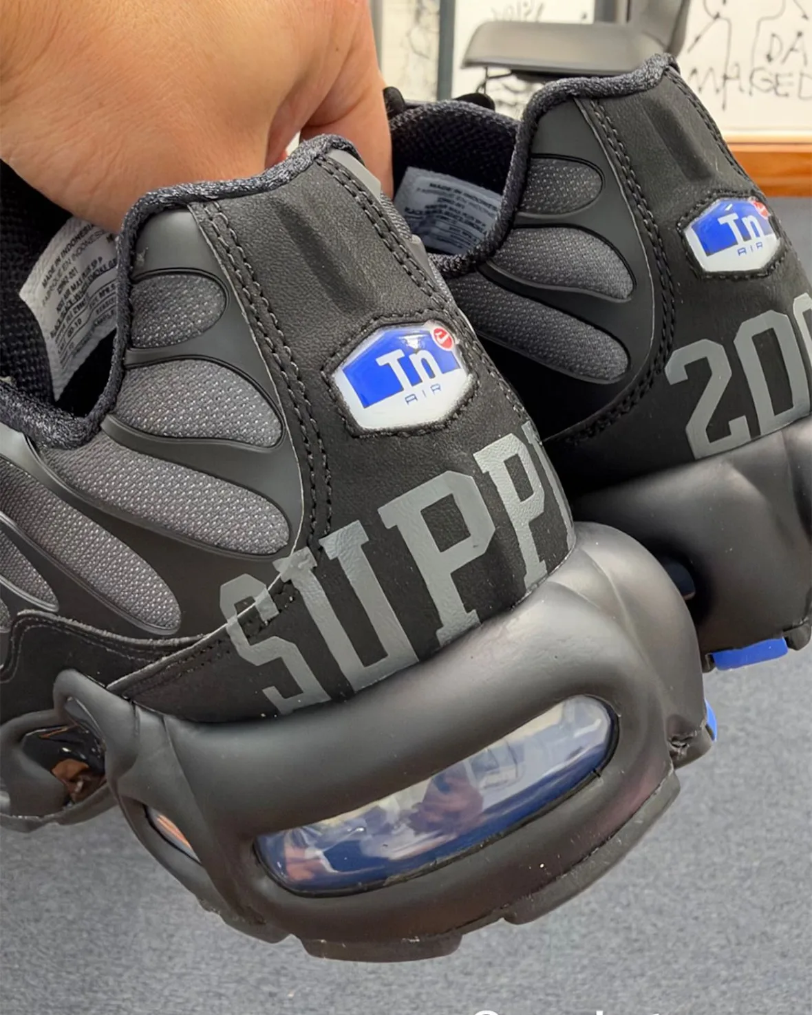 Heel view held in hand: black pair, grey SUPPLY and 200 lettering, blue Tn badges, clear heel air unit, Plus x Supply