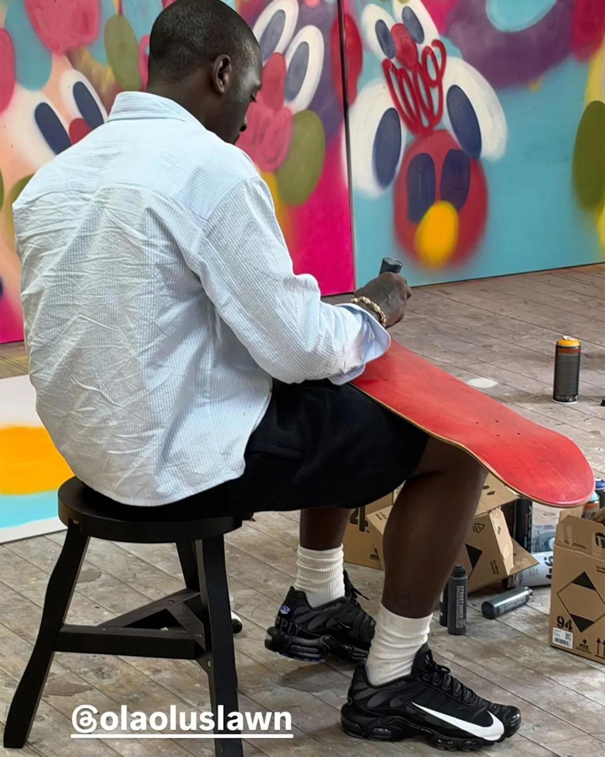 Seated person painting a red skateboard deck in a colourful studio, black pair with white swoosh on feet, Plus x Supply