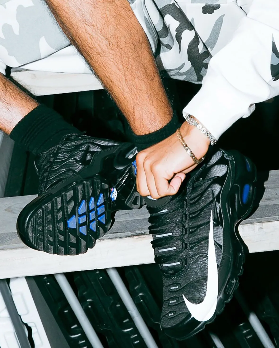 On-foot detail of black pair, blue outsole studs visible, big white swoosh, camo shorts and white sleeve, Plus x Supply