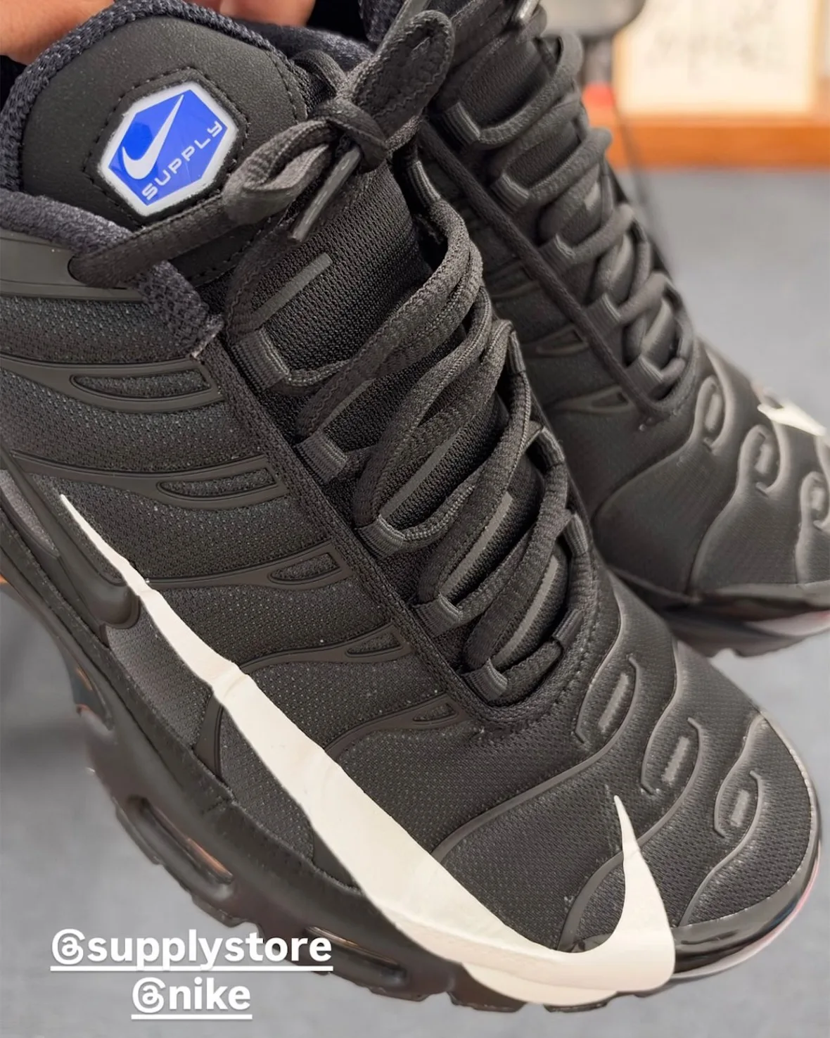 Top-down in-hand view of black pair with oversized white swoosh and blue Supply hexagon tag on tongue, Plus x Supply