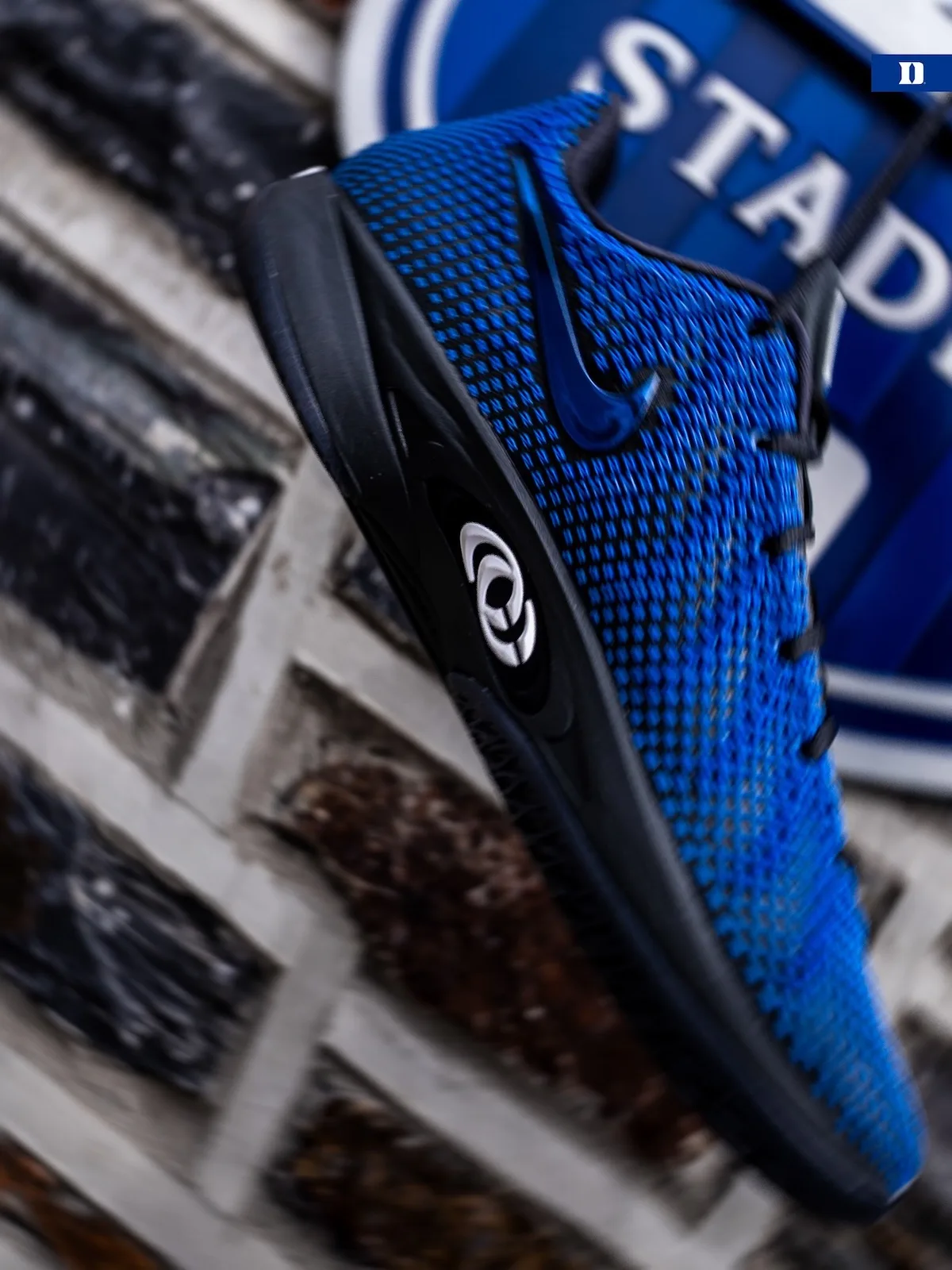 Angled lateral view, blue knit, black sole, white circular logo, blurred blue stadium sign, Duke