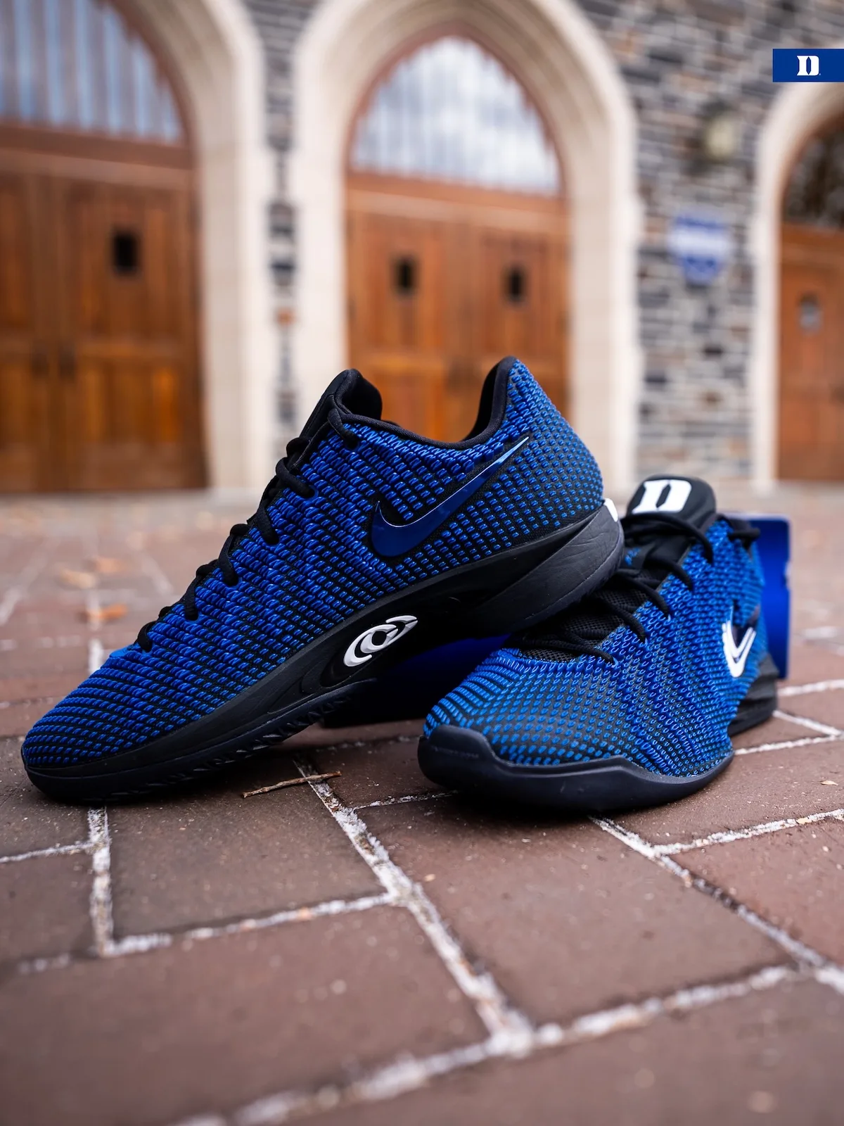 Pair on brick paving, blue knit, black soles, white Duke D tongue, arched doors behind, Duke