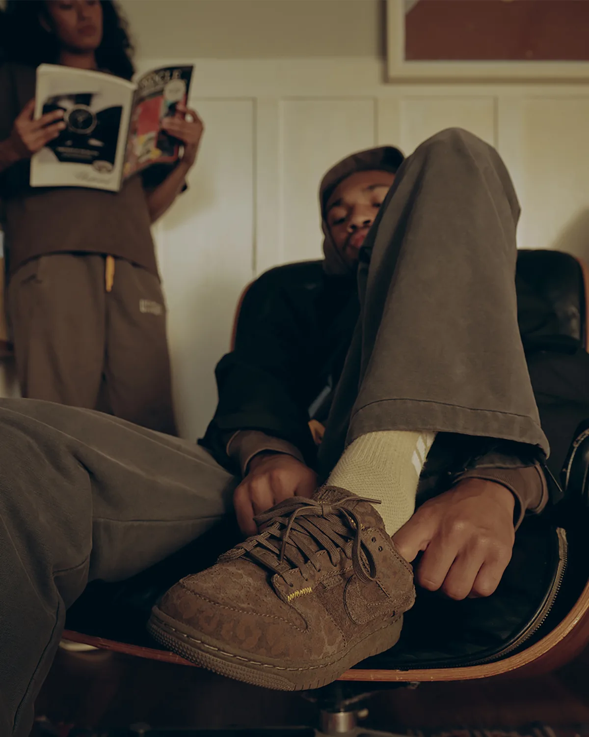 Low-angle on-foot shot of brown leopard-textured suede low-top, person tying laces, person reading behind, Natural Selection
