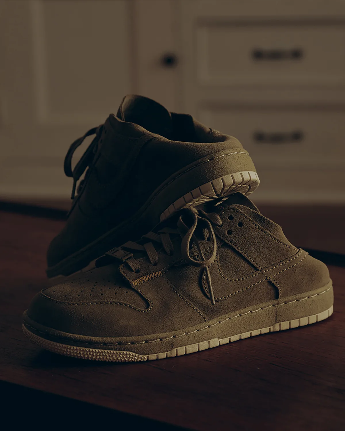 One olive suede low-top stacked on the other, cream sole, cabinet doors behind, Natural Selection