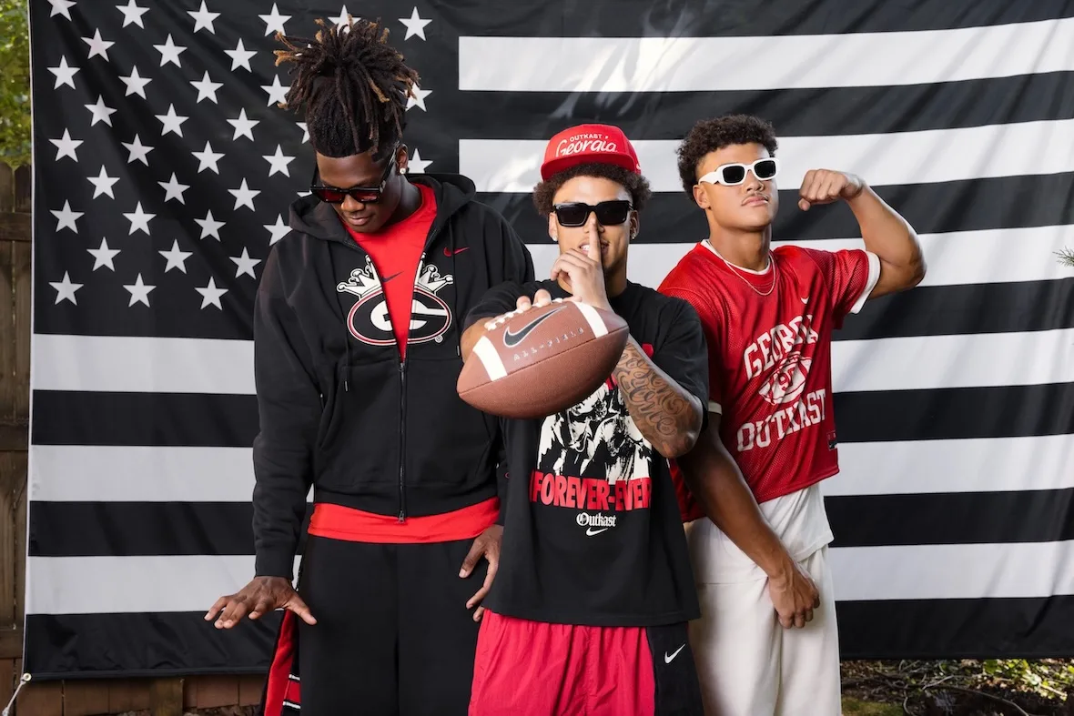 Three people before a black and white flag, one holding a Nike football, Georgia and Outkast tops, Outkast Football