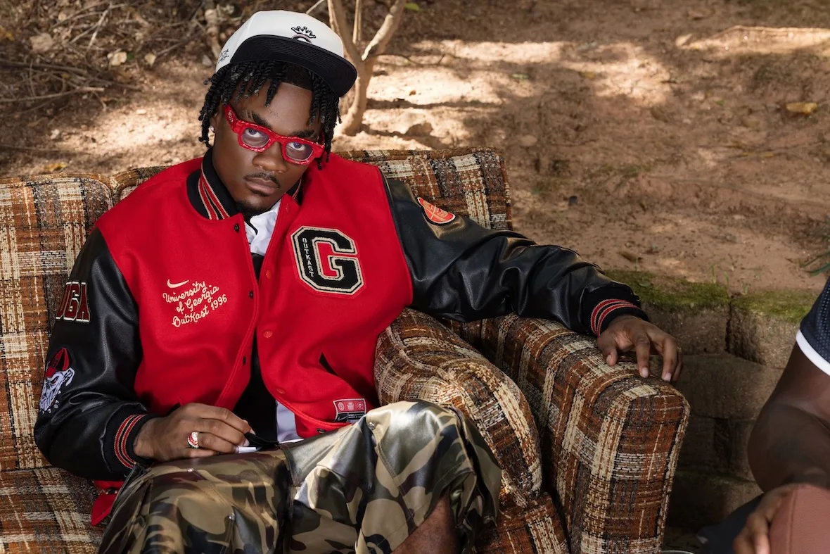 Seated on-foot lifestyle shot in red varsity jacket with G patch, camo pants, plaid armchair, Outkast Football Collection
