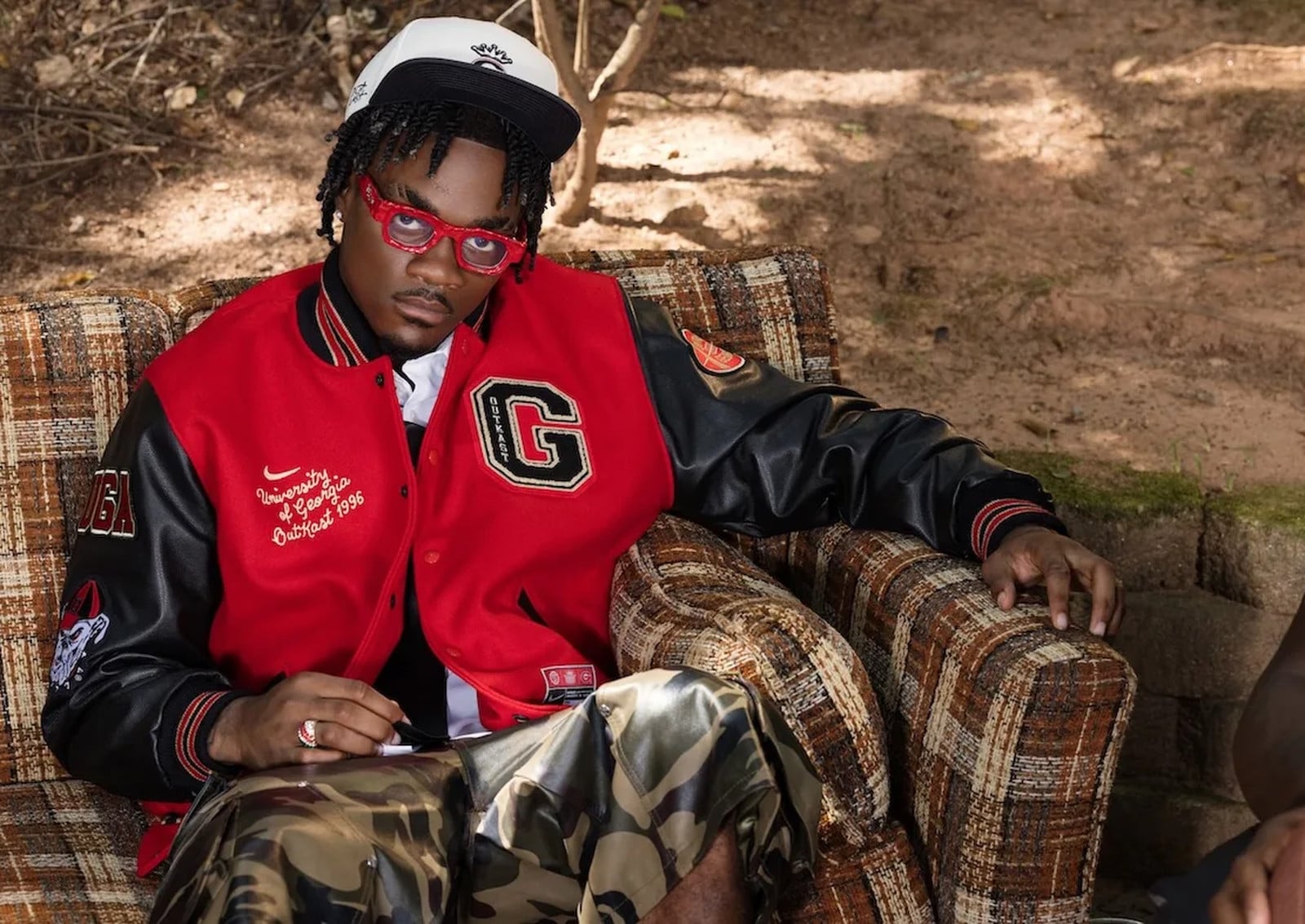Person seated on plaid sofa in red and black varsity jacket, red glasses, camo, Nike x Georgia x Outkast Football Collection
