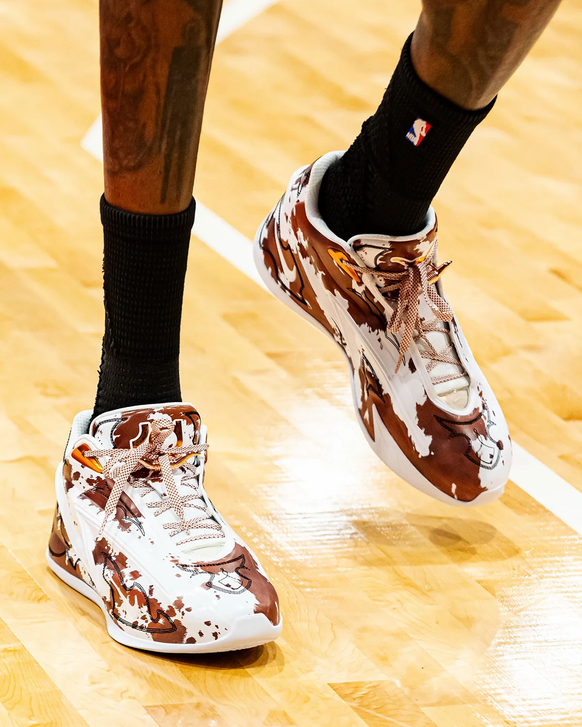 On-foot top-down angled view, brown and white cow-print upper, pink woven laces, black NBA socks, Texas