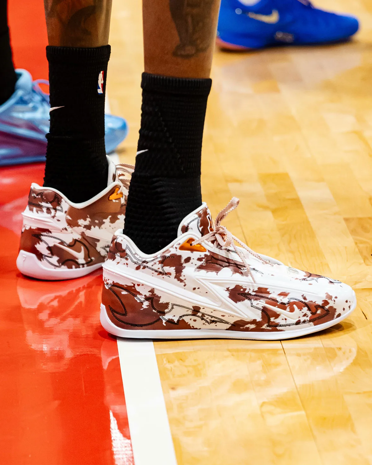 On-foot pair on court, brown and white cow-print upper, orange tabs, black NBA socks, blue shoe behind, Texas