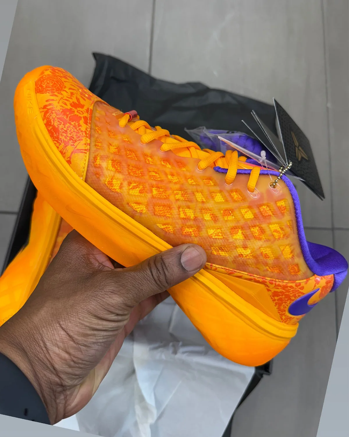 Lateral view in hand: orange upper, purple heel swoosh and collar, black hangtag, box behind, Mamba Eterno