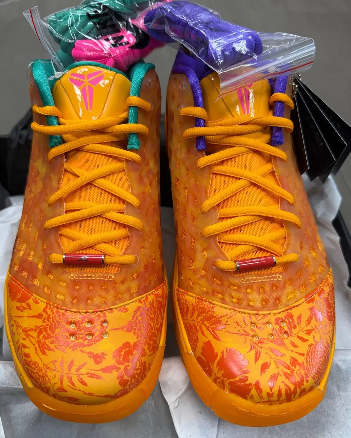 Top-down pair in box: orange floral toes, teal and purple collars, red lace tips, spare laces in bags, Mamba Eterno