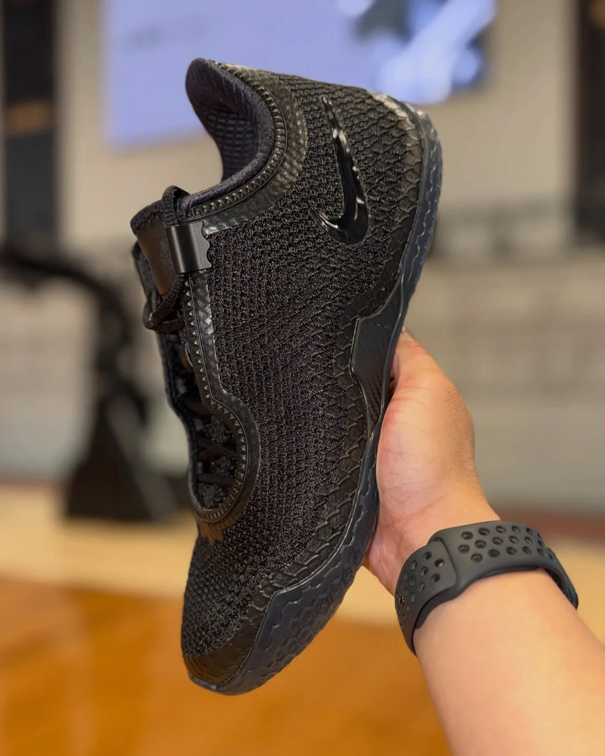 In-hand view held up by a wrist with black sport band, black mesh upper and glossy swoosh, Black