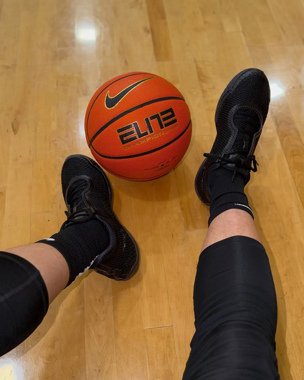 Top-down on-foot view of black pair on a wood court beside an orange Nike Elite basketball, Black